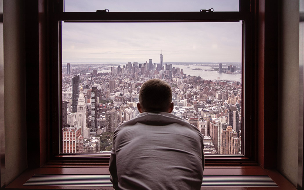 Solitude : Un homme regarde par la fenêtre l'horizon de la ville. / Solitude: Man looking out a window at the city skyline